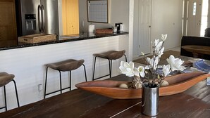 Moderni keittiö, jossa on ruostumattomasta teräksestä valmistettu jääkaappi, puiset baarituolit ja puinen ruokapöytä, jonka päällä on maljakko valkoisia kukkia.