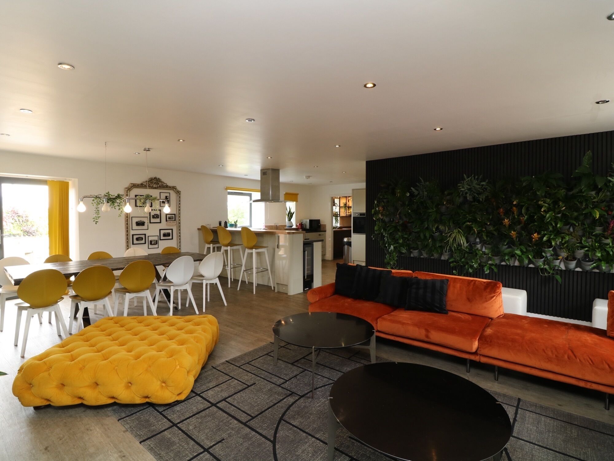 Un salon moderne avec un canapé, un pouf et une table basse, un coin repas avec des chaises et une table, et une cuisine en arrière-plan.