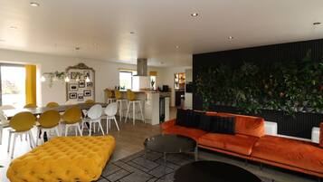 Un salon moderne avec un canapé, un pouf et une table basse, un coin repas avec des chaises et une table, et une cuisine en arrière-plan.