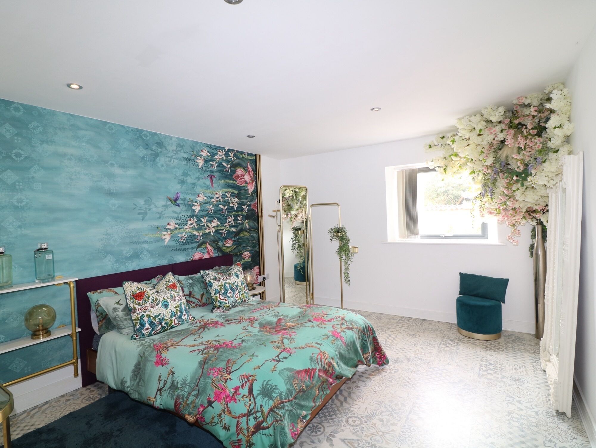 A bedroom with a bed, a large mirror, a window with flowers, and a patterned floor.