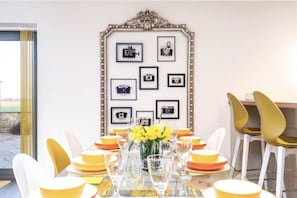 Un coin repas avec une table dressée pour quatre personnes, entourée de chaises jaunes, d’un miroir décoratif orné de photos encadrées et d’un vase rempli de fleurs jaunes.
