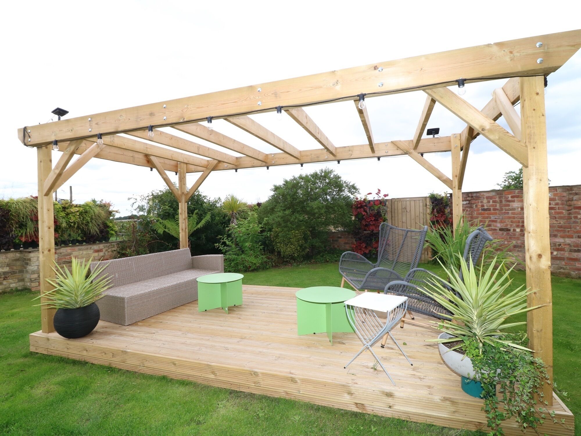 Una pérgola de madera con un sofá integrado y dos pequeñas mesas, rodeada de plantas en macetas y un muro de ladrillo.