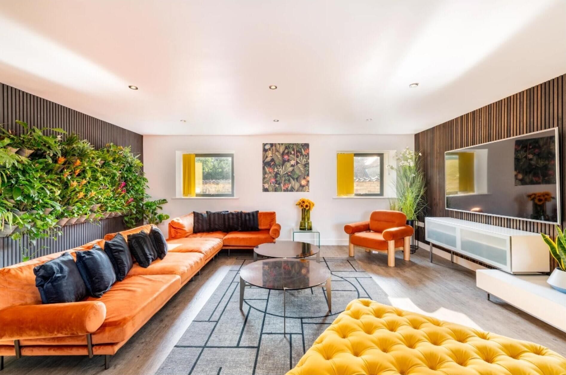Un salon moderne avec un canapé, des fauteuils, une table basse et un téléviseur. Des plantes et des œuvres d’art décorent les murs.