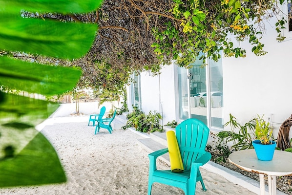 A covered outdoor seating area with turquoise chairs and a small table, surrounded by greenery and a white building.