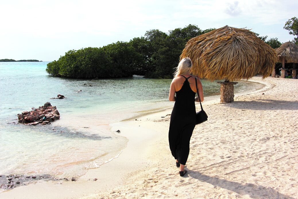 Een strand met helder turquoise water, een vrouw in een zwarte jurk en rieten parasols.