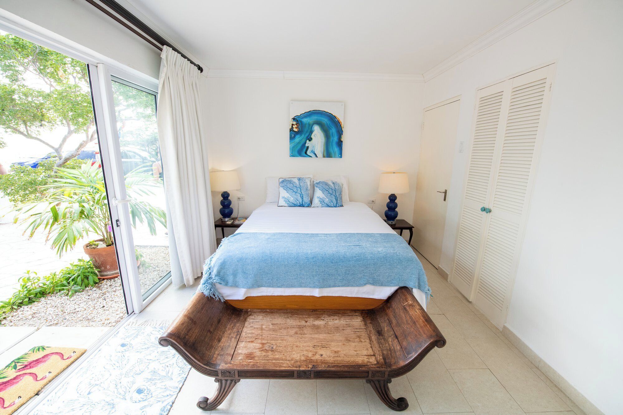 Une chambre à coucher comprenant un lit, deux tables de chevet, une commode, un banc en bois, une fenêtre avec des rideaux, une porte et un tableau accroché au mur.
