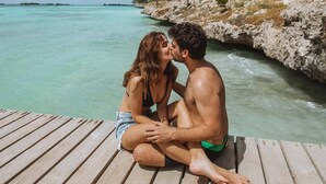 A couple sitting on a wooden dock by a turquoise sea, with rocky terrain and sparse vegetation in the background.