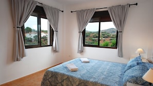 A bedroom with two windows, a bed with a patterned bedspread, and a nightstand with a lamp.