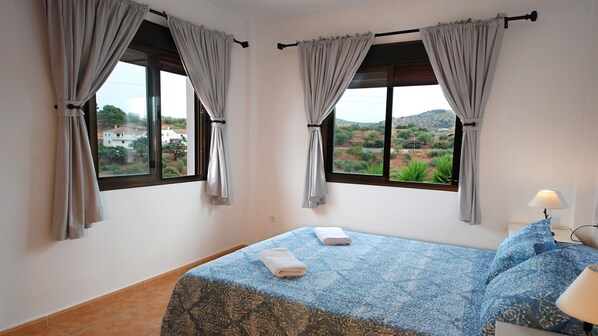 A bedroom with two windows, a bed with a patterned bedspread, and a nightstand with a lamp.