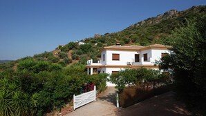 A two-story house with a tiled roof, surrounded by greenery and hills.