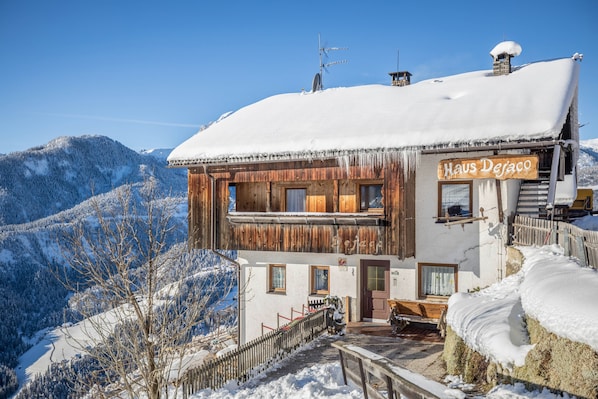 A wooden chalet with a sign reading 'Haus Deraco' amidst a snowy mountainous landscape.