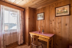 Un interior de madera con una mesa, una silla, cortinas de encaje y cuadros en las paredes.
