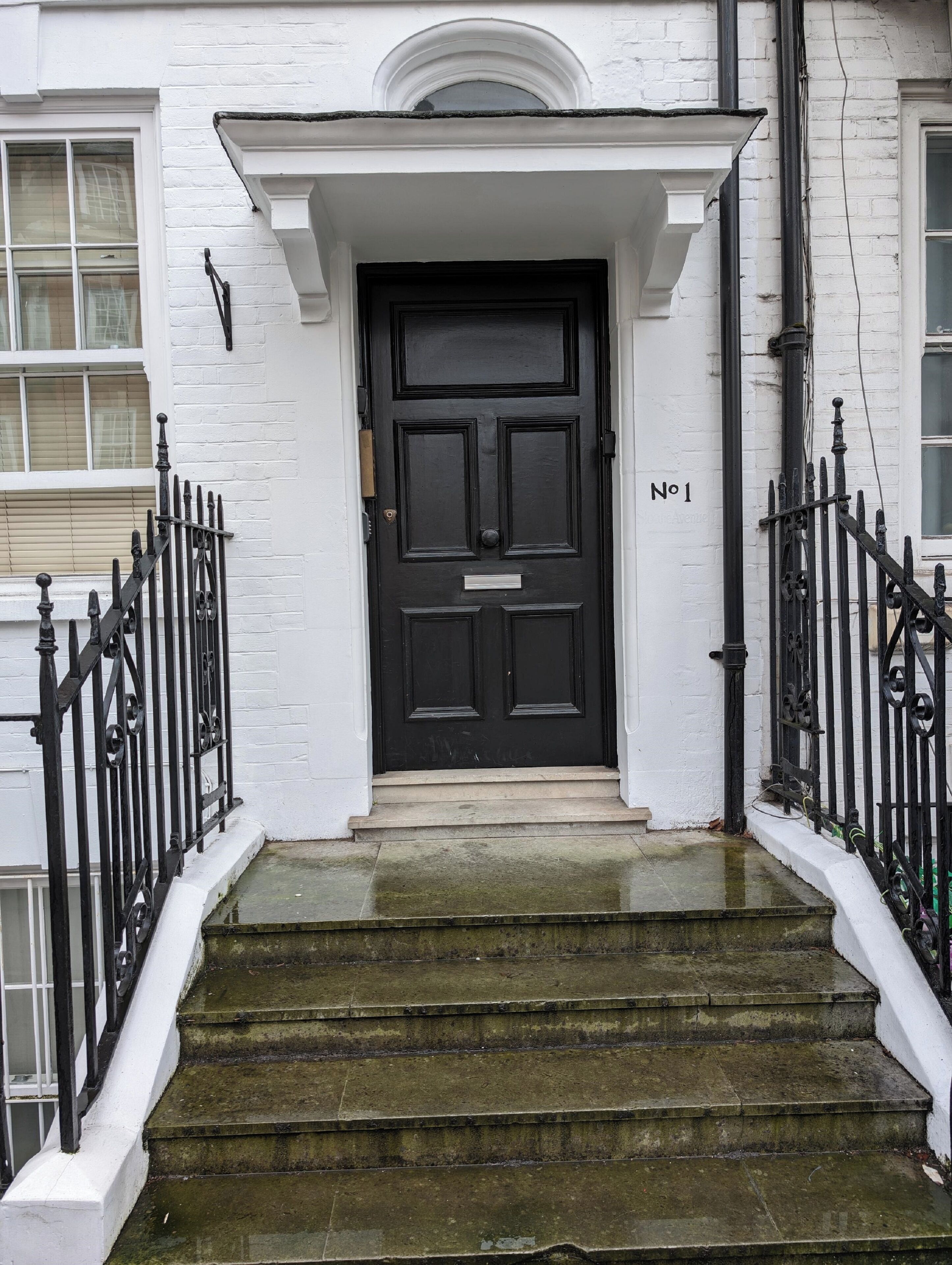 A black door with a white number plaque, flanked by black iron railings and a white wall.