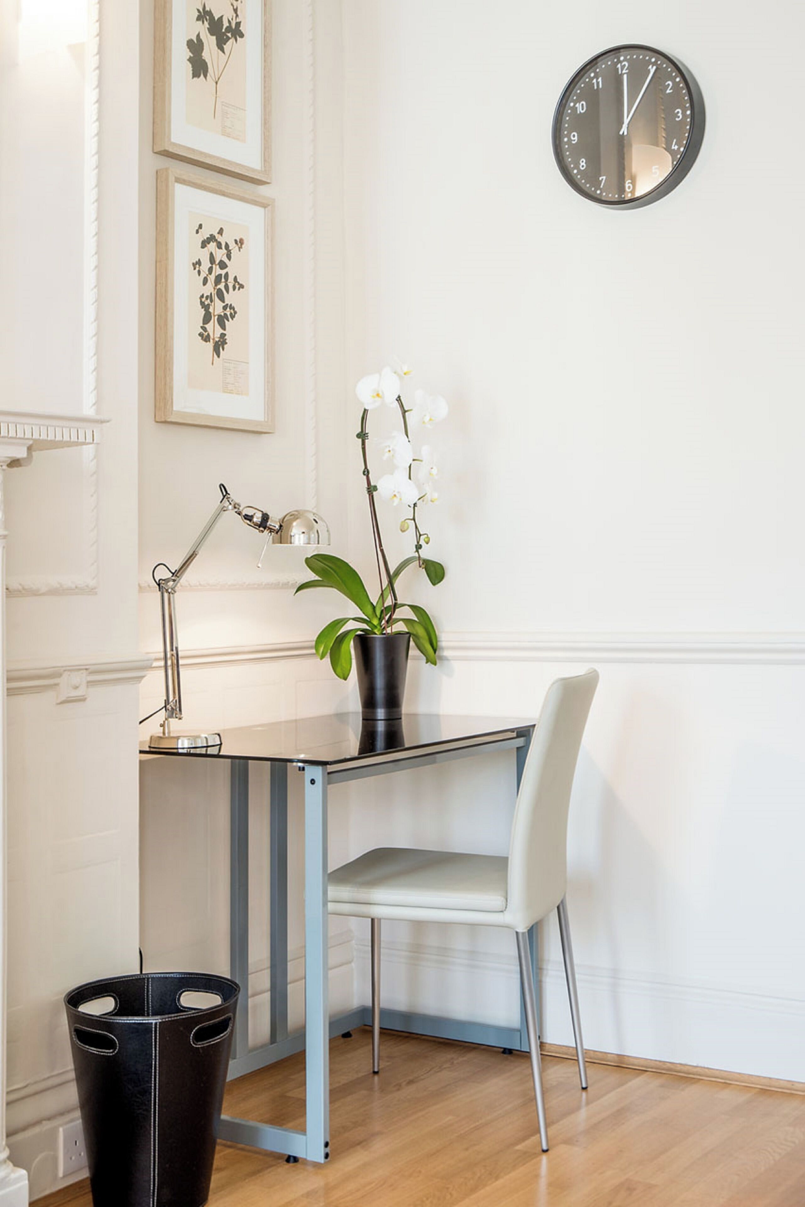 Un escriptori modern amb una làmpada, una orquídia en test i una cadira en una habitació ben il·luminada, amb gravats botànics emmarcades a la paret.