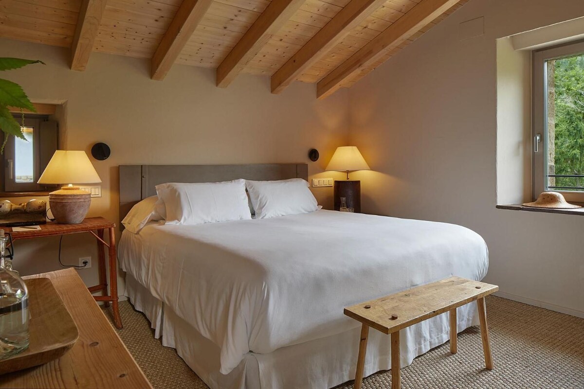 A bedroom with a bed, bedside lamps, a wooden bench, a window, and a view of greenery.