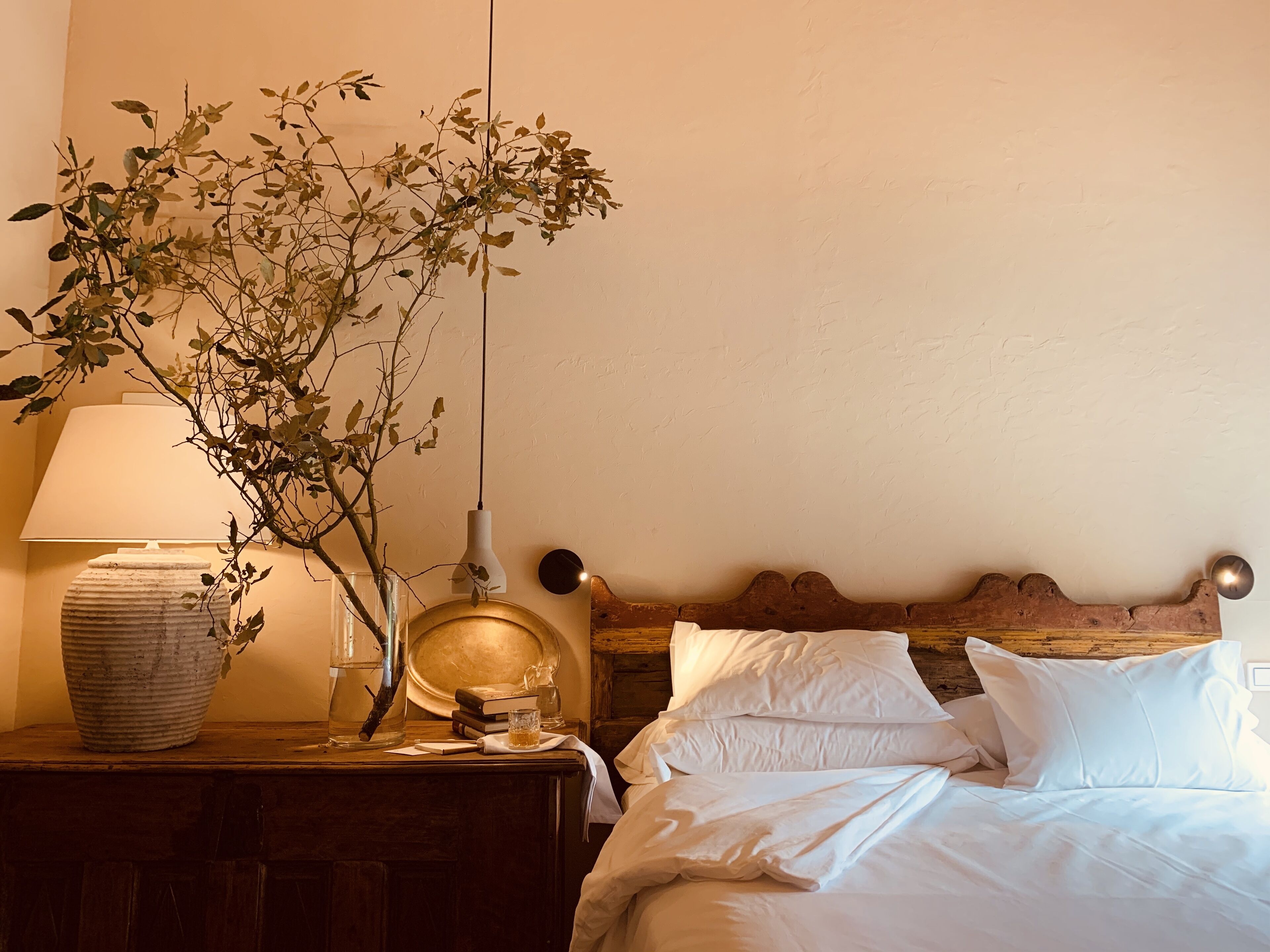 A neatly made bed with white linens, a bedside lamp, a vase with a branch, and a round mirror on a wooden nightstand.