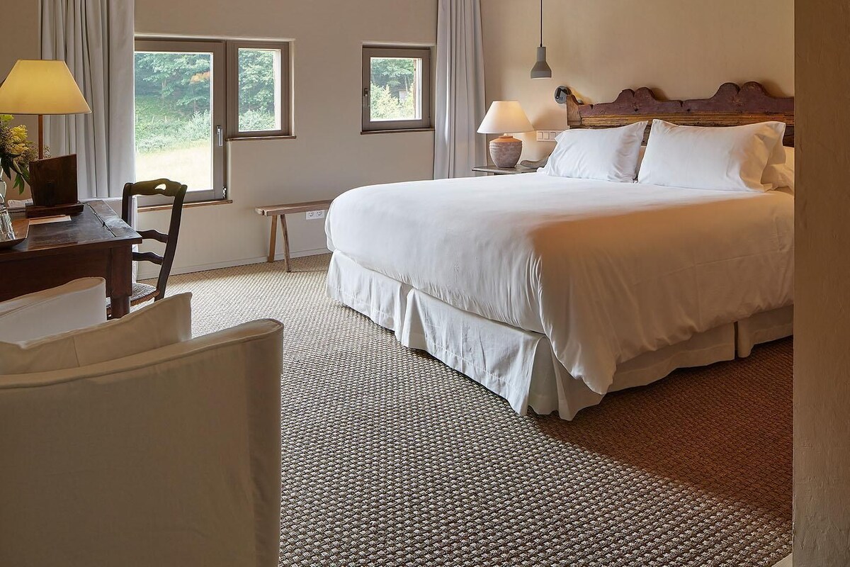 A bedroom with a large bed, two bedside lamps, a wooden headboard, a desk with a chair, and a window with curtains.