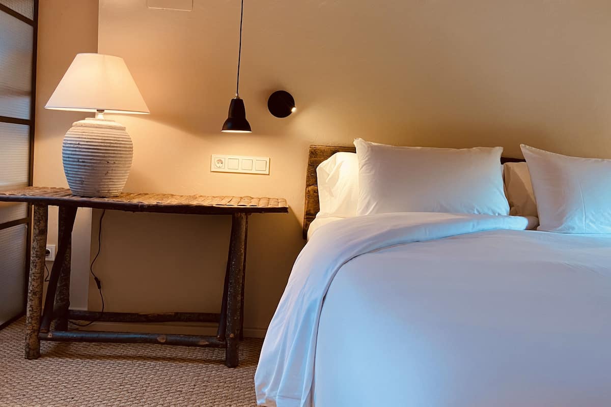 A neatly made bed with white linens, a bedside table with a lamp, and a wall-mounted light fixture.