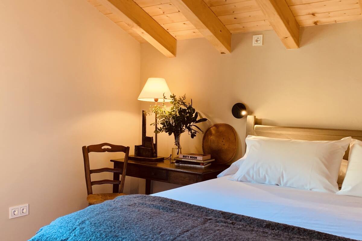 A bedroom with a bed, a desk, a chair, a lamp, and a bedside table.