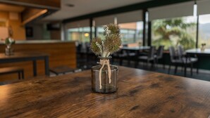 Une table en bois sur laquelle est posé un vase en verre transparent contenant une plante. L’ensemble semble représenter un espace intérieur moderne, avec un comptoir en bois et des chaises en arrière-plan.