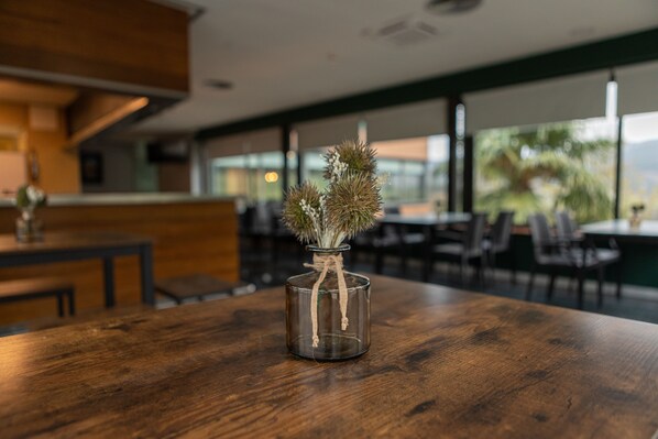 Uma mesa de madeira com um vaso de vidro transparente contendo uma planta. O ambiente parece ser um espaço interior moderno, com um balcão de madeira e cadeiras ao fundo.