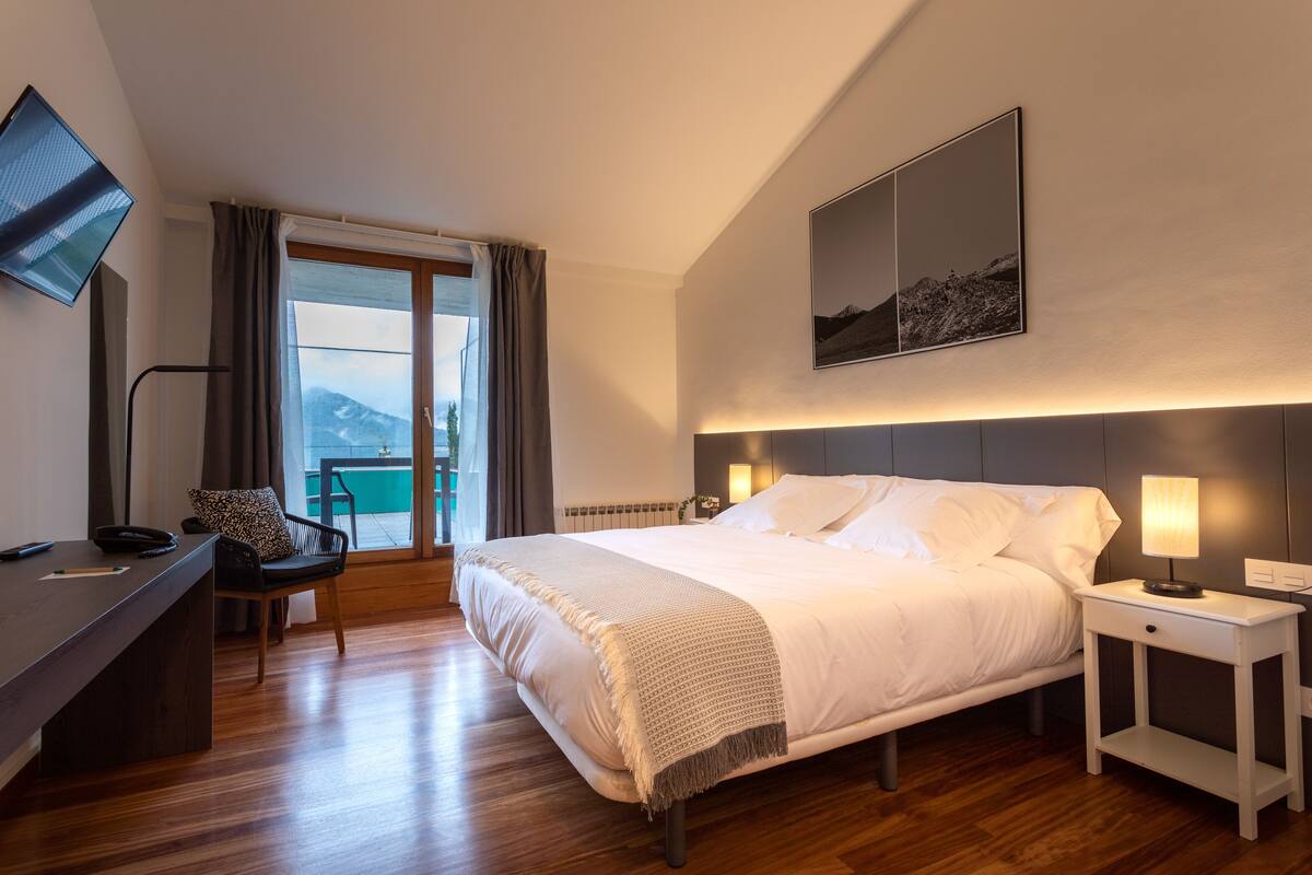 Hotelkamer met een bed, nachtkastjes, een bureau met stoel, een televisie en een groot raam met gordijnen.