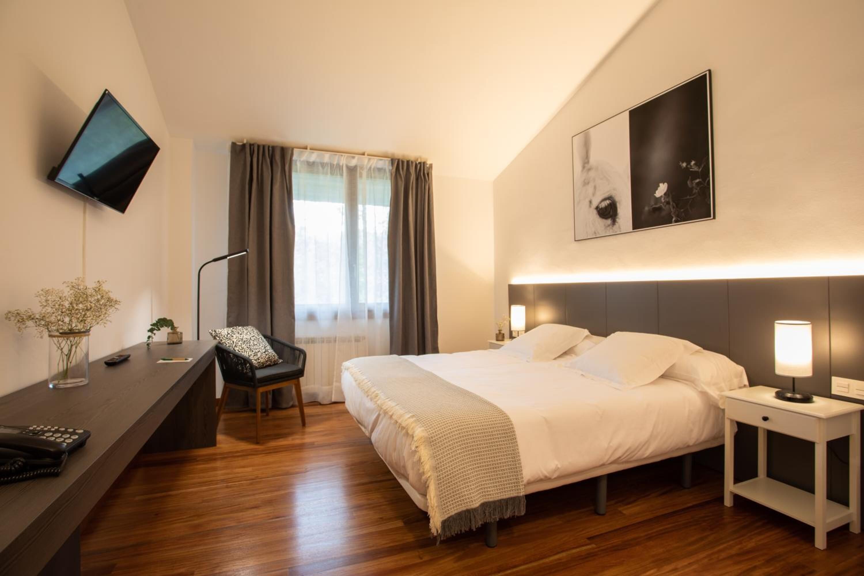 A modern hotel room with a bed, bedside tables, a desk, a chair, a TV, and a window with curtains.