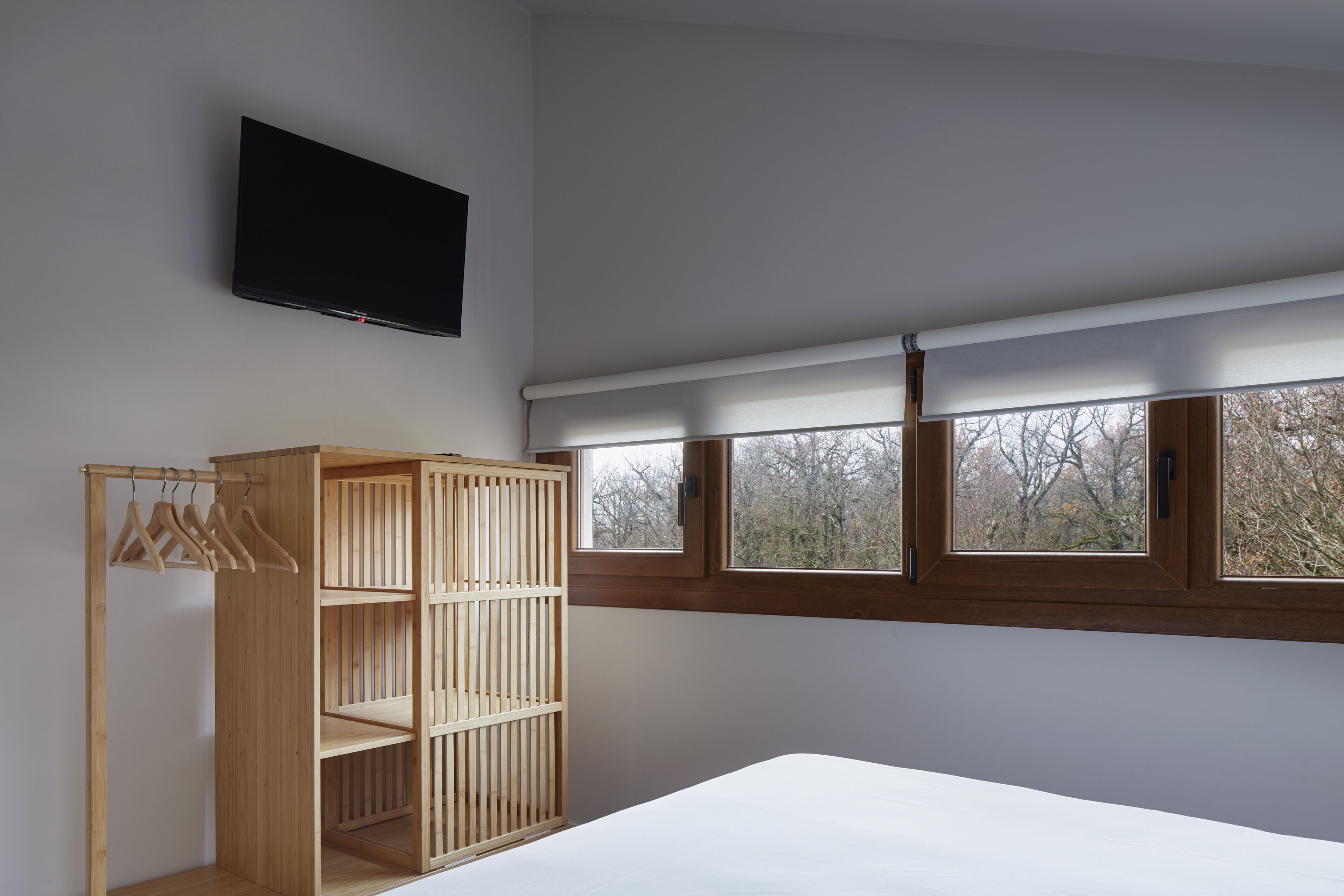 A bedroom with a bed, a wooden wardrobe, a TV mounted on the wall, and a window with a view of trees.