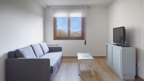 A modern living room with a grey sofa, a small white table, a television on a cabinet, and a window with blinds. - Aritzalko (Etxarri-Aranatz)
