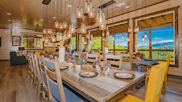 A wooden interior with a long dining table, hanging light fixtures, and large windows with a view of greenery. - Friendship Mtn Retreat: : Indoor Pool, Mtn View, Theater, Games, Hot Tub, Wifi (Pigeon Forge)