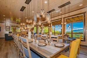 A wooden interior with a long dining table, hanging light fixtures, and large windows with a view of greenery.