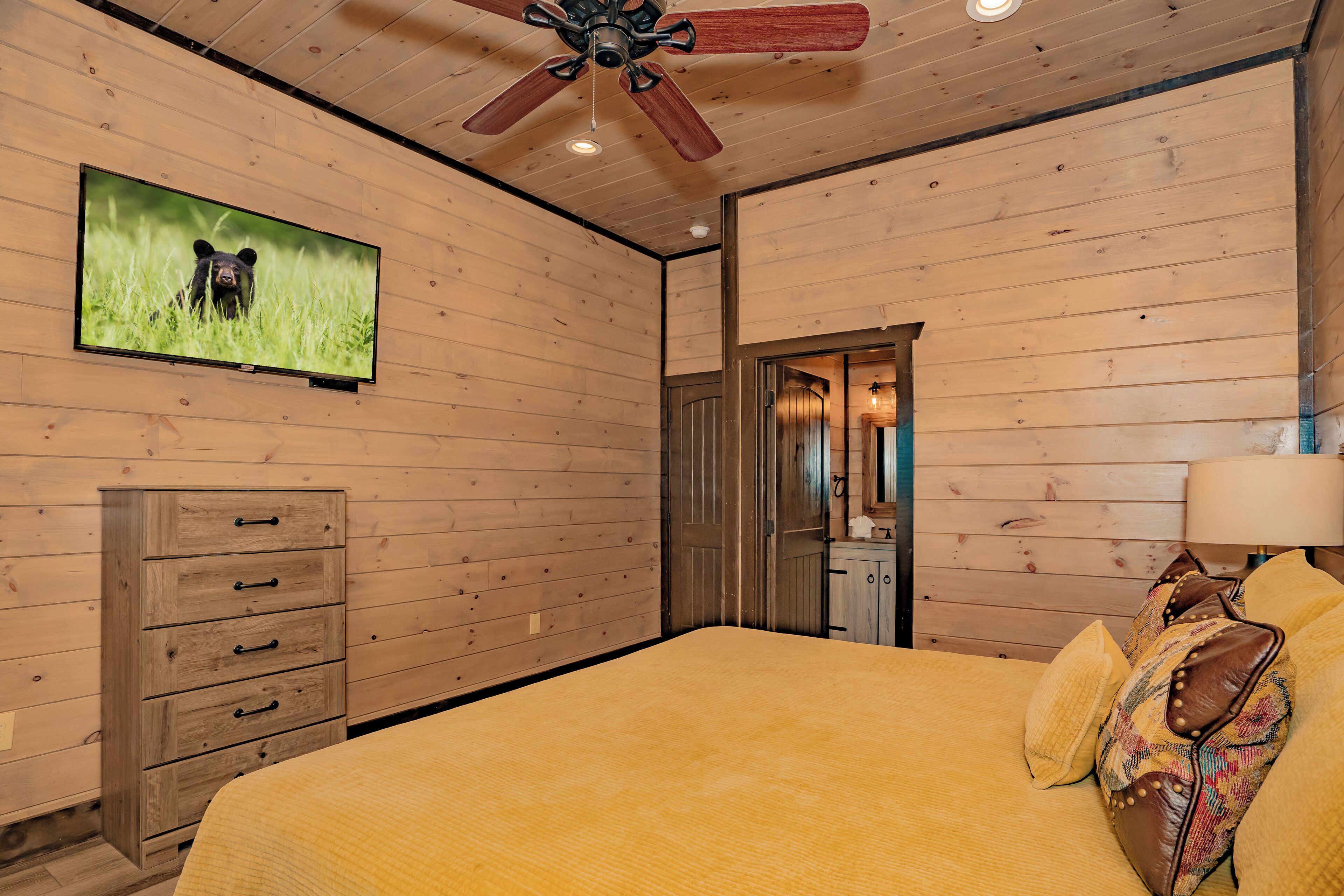 A bedroom with wooden walls, a bed with yellow bedding, a TV on the wall, a wooden dresser, and a ceiling fan.