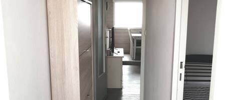 A narrow hallway with a wooden floor, a white door on the right, and a bed with striped bedding in the background.