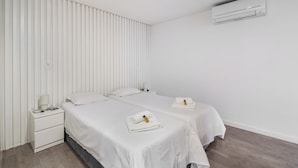 Ett hotellrum med två sängar, ett nattduksbord med en lampa, en väggmonterad luftkonditioneringsanläggning och ett fönster med persienner.