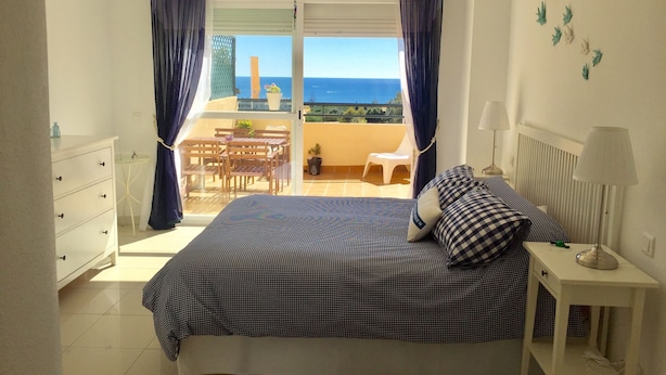 A bedroom with a bed, a bedside table with a lamp, a dresser, and a balcony with a table and chairs. - Townhouse with sea view walking distance to the beach - 1 (Mijas Costa)