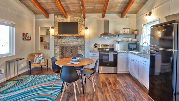 A modern kitchen with a stone fireplace, wooden beams, a dining area with a colorful rug, and a large refrigerator. - Quaint Cottages in Canyon Lake! Perfect for kids! (Canyon Lake)