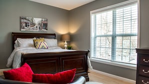 Une chambre avec une grande fenêtre, un lit recouvert d’une literie à motifs et un oreiller rouge.
