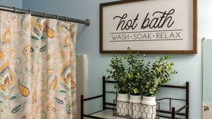 Une salle de bain avec un rideau de douche, un panneau décoratif encadré au-dessus d’une étagère ornée de plantes et une baignoire blanche.