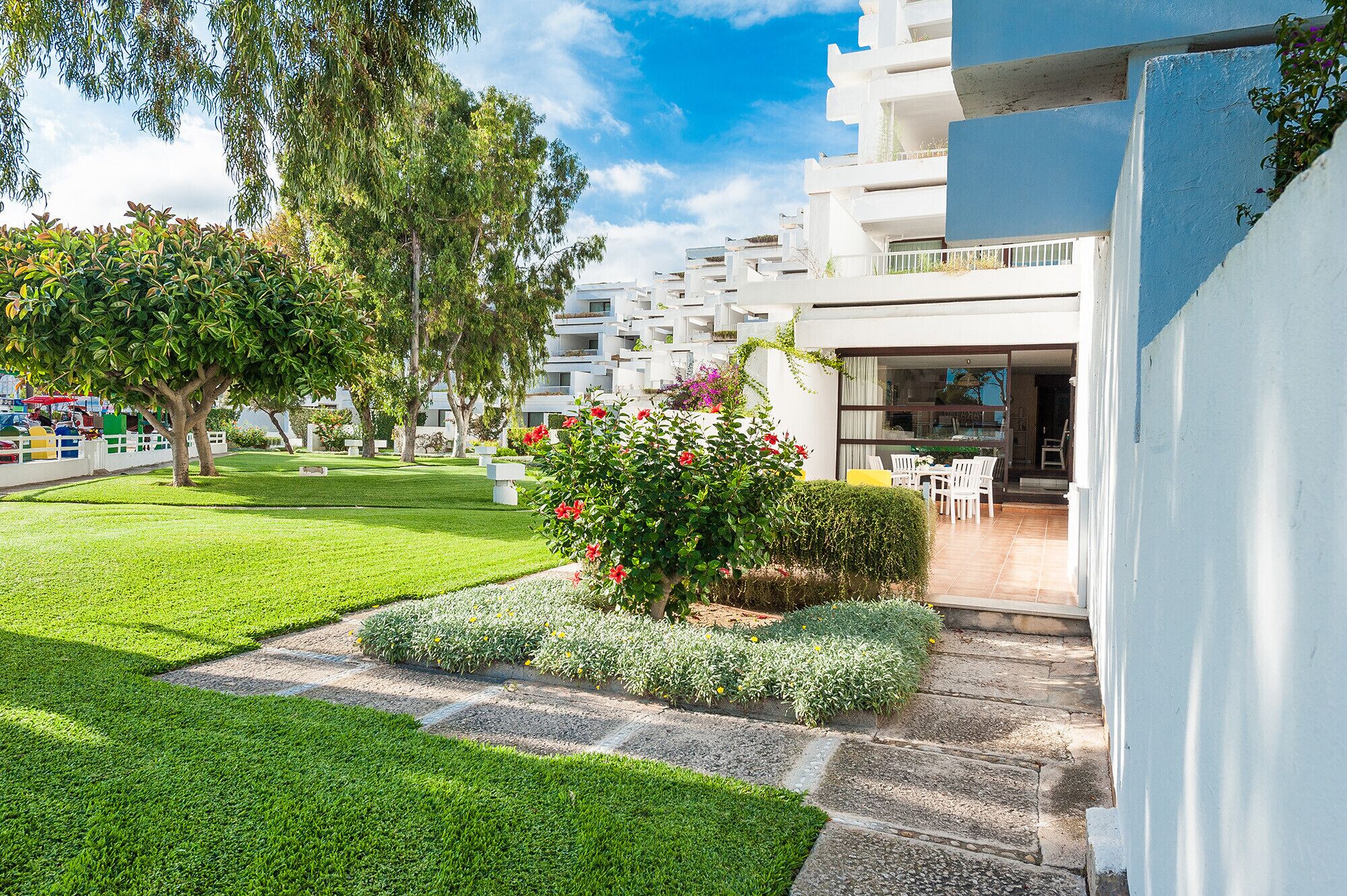 Un edificio moderno con fachada blanca, un jardín bien cuidado con césped verde y plantas en flor, y un patio con muebles de exterior.
