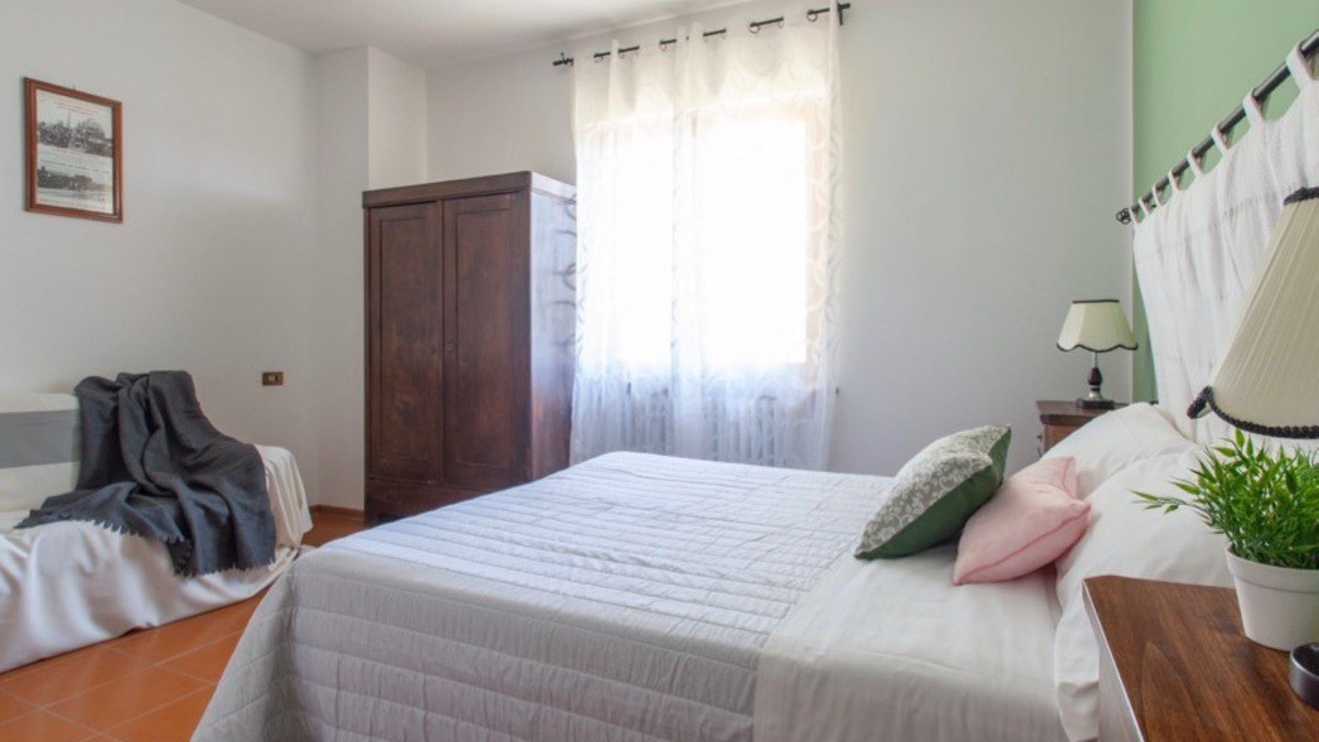 Un dormitorio con una cama, una mesita de noche, una lámpara, una planta en maceta y un armario.