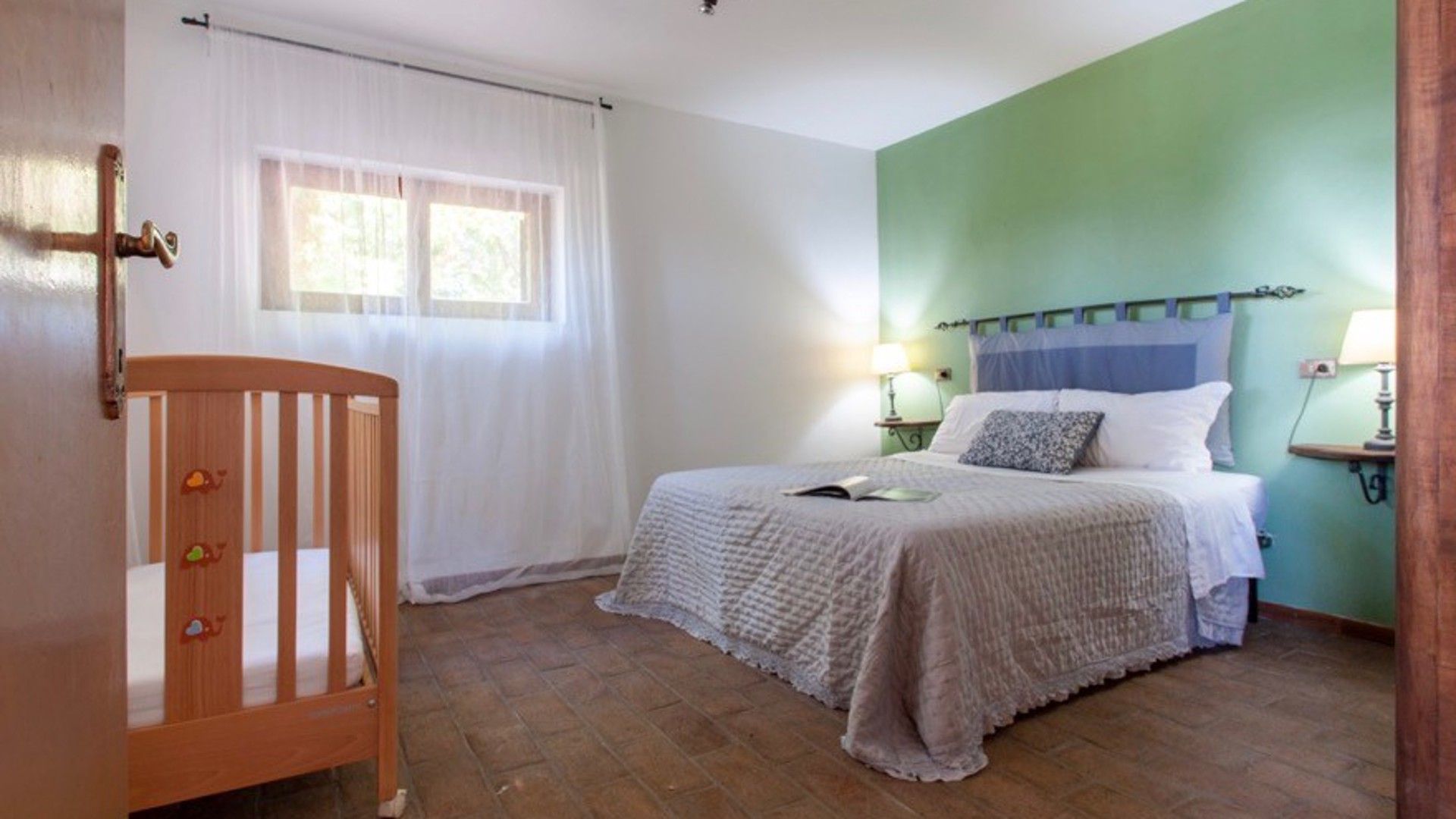 A bedroom with a single bed, a wooden crib, a window with white curtains, and a green wall.