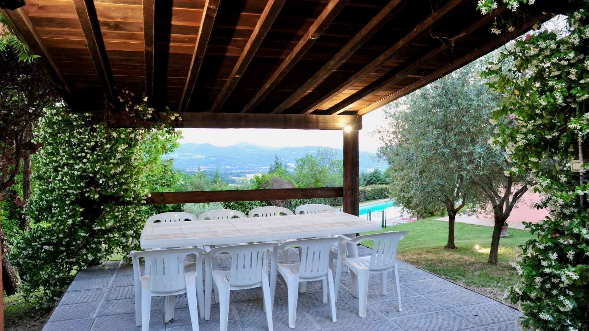 Un patio al aire libre con una mesa de madera y sillas de plástico blanco, rodeado de vegetación y vistas a montañas distantes.