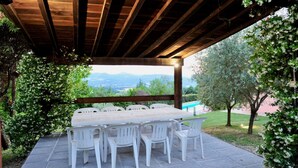 Un patio al aire libre con una mesa de madera y sillas de plástico blanco, rodeado de vegetación y vistas a montañas distantes.