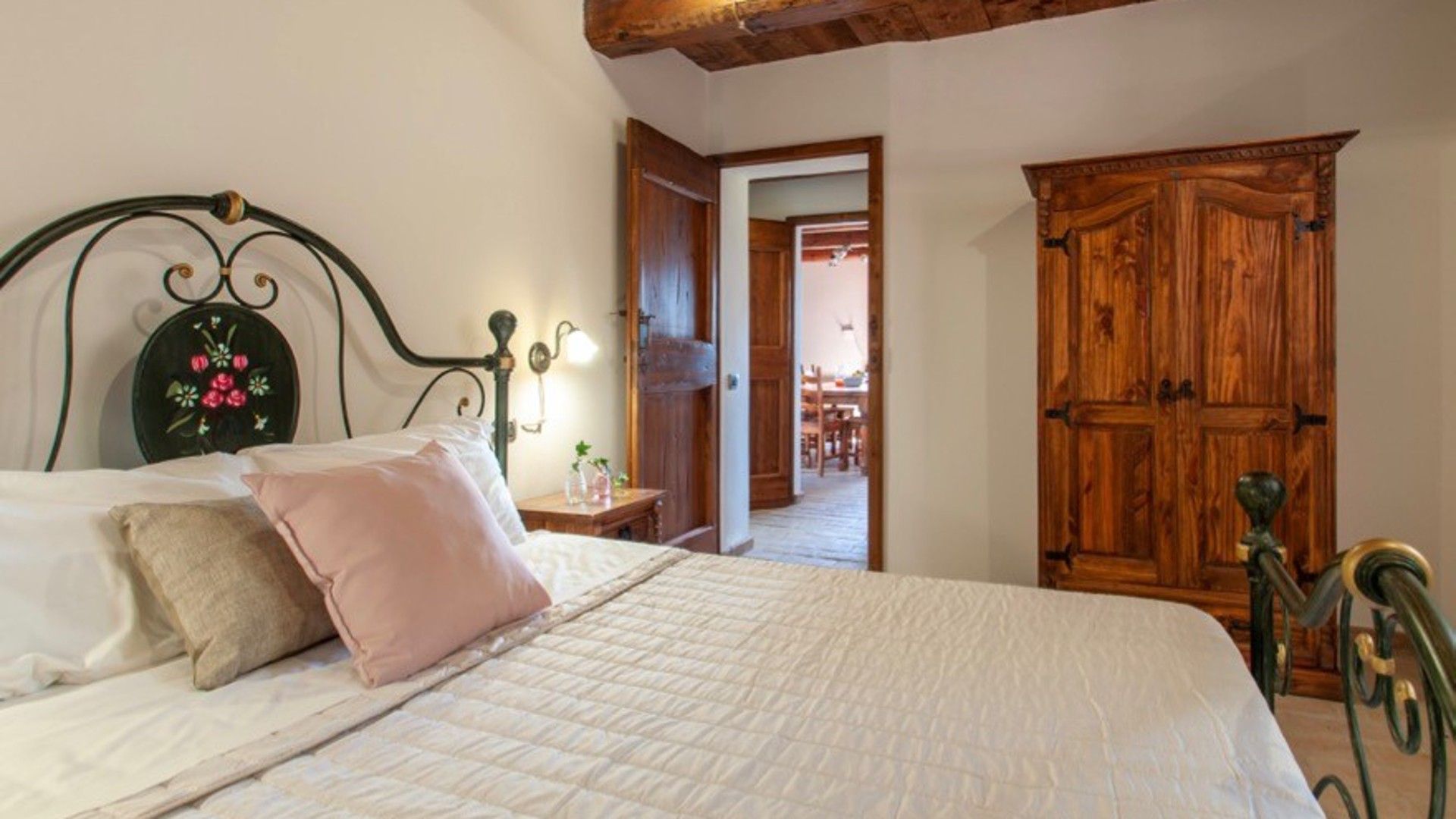 A bedroom with a bed, a nightstand, a wooden wardrobe, and a door leading to another room.