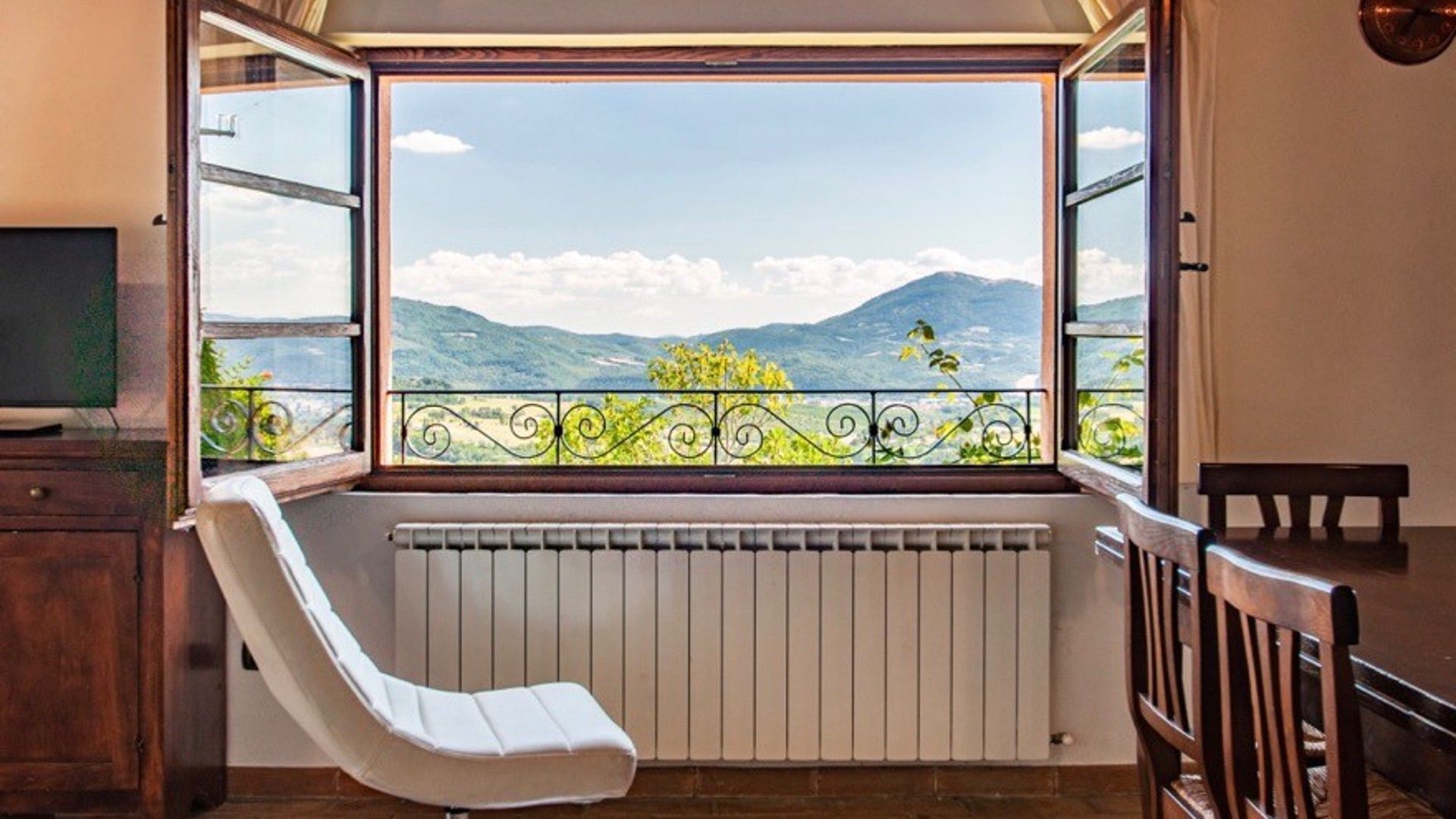 Een kamer met een groot raam, een witte stoel, een radiator en een houten tafel.