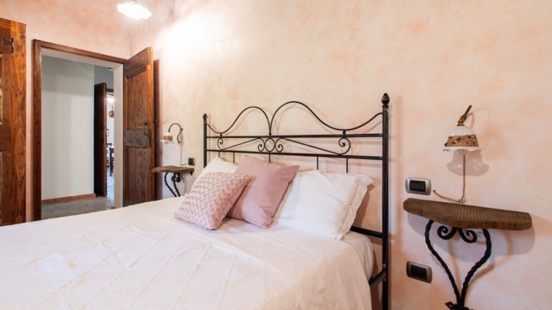 A neatly made bed with a metal headboard, a bedside table with a lamp, and a wall-mounted alarm clock.