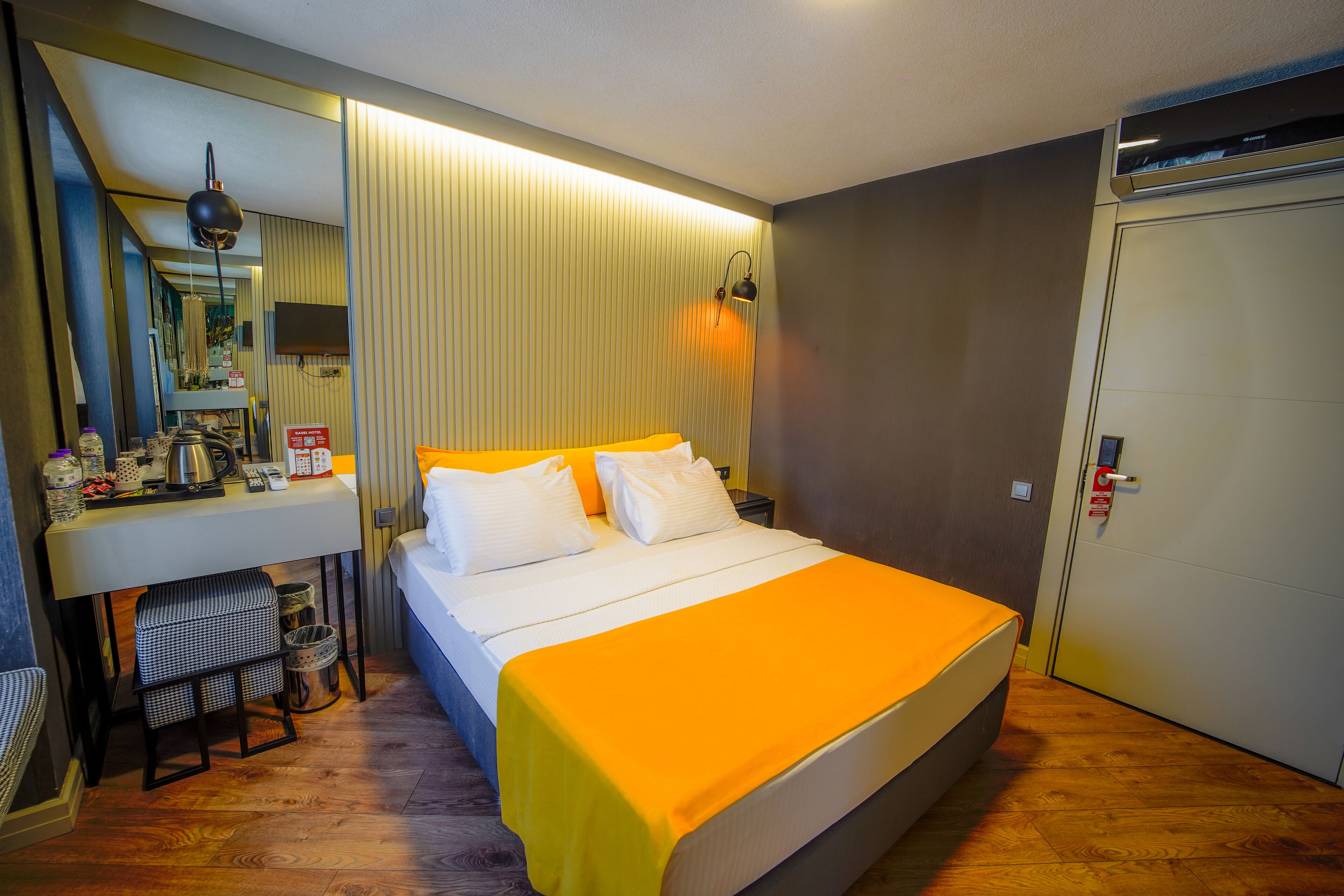 Quarto de hotel com cama, escrivaninha com espelho, pequeno espaço para kitchenette e guarda-roupa.