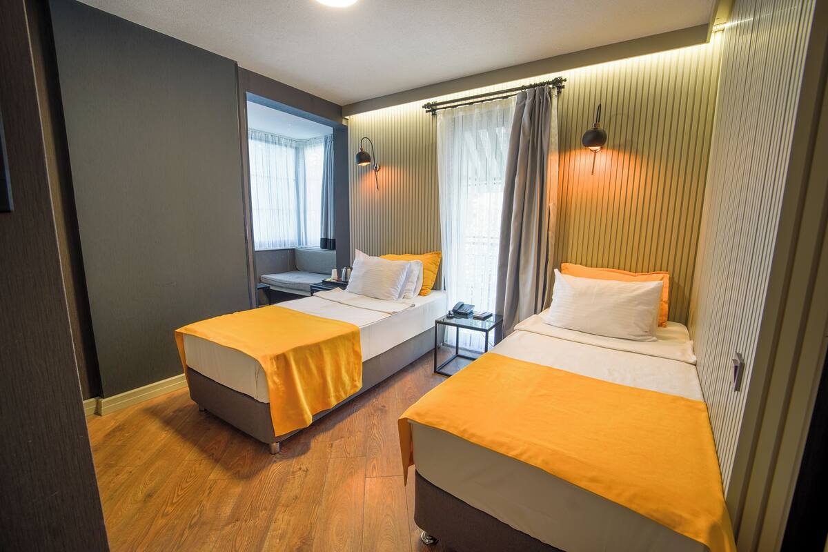 Habitación de hotel con dos camas individuales, una mesita, una silla, una ventana con cortinas y una lámpara de noche.