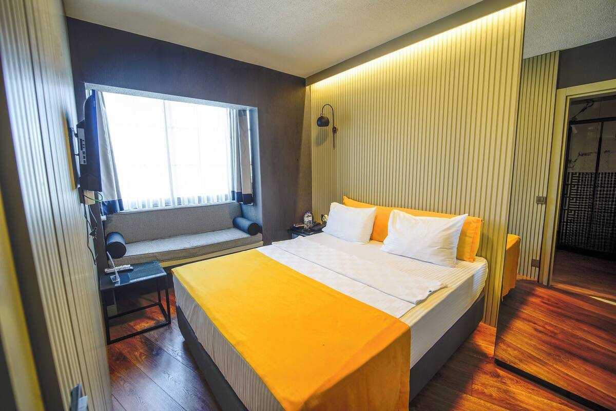 Una habitación de hotel con una cama, una ventana, una mesita de noche y una lámpara de pared.