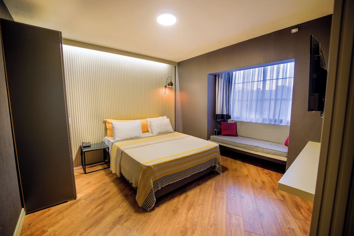 Habitación de hotel moderna con una cama grande, una mesita de noche, una pequeña mesa, un banco junto a la ventana y un televisor montado en la pared.